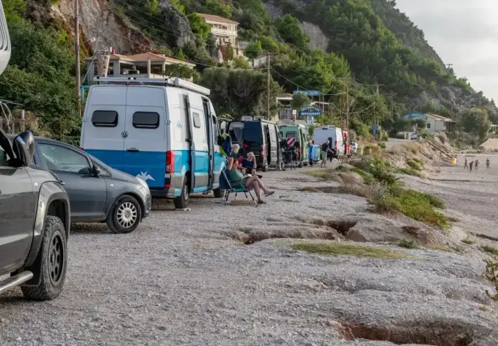 Une rangée de camping-cars en Grèce, certains garés sur un chemin de gravier près d'une plage. Des gens se détendent à l'extérieur.