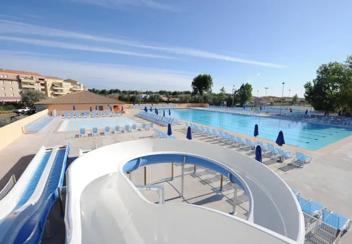 Piscine et toboggans aquatiques dans une aire de camping-car au Grau-du-Roi. Des chaises longues bleues et des parasols sont disposés autour des bassins.