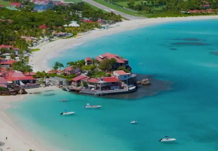 Vue aérienne de Saint-Barthélemy, avec ses plages de sable blanc, ses eaux turquoise et ses villas aux toits rouges. Un voilier navigue paisiblement.