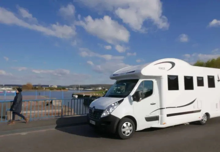 Un camping-car blanc roule sur un pont, avec une personne marchant à côté. Le ciel est bleu avec des nuages. Idéal pour la consommation camping-car.