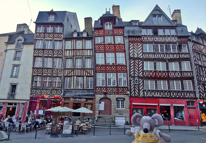 Une souris en peluche pose devant des maisons à colombages colorées, un aperçu de ce que faire à Rennes.