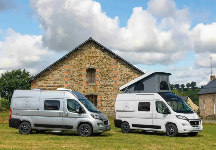Deux camping-cars, un argenté et un blanc avec toit relevable, devant une bâtisse en pierre. Idéal pour savoir comment choisir son camping-car.