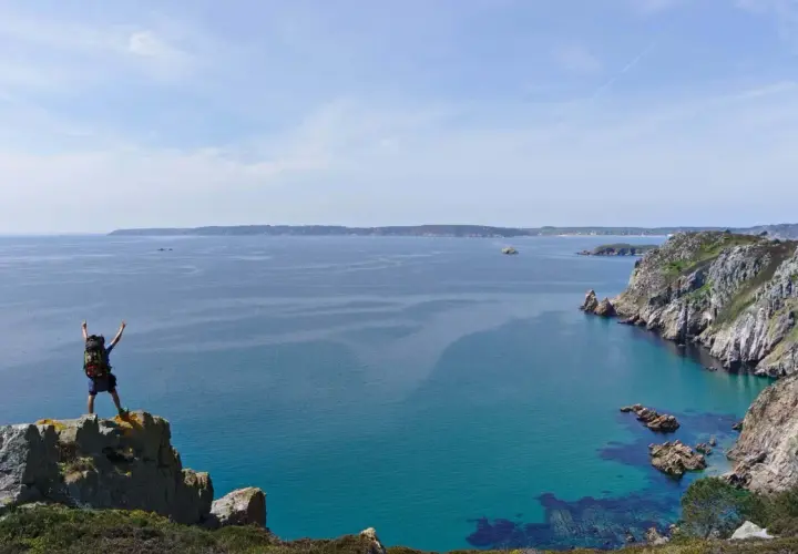 Randonneur victorieux au sommet d'un rocher, bras levés, dominant la mer turquoise et les falaises de GR34 Crozon.