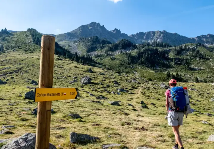 Randonneuse sur le GR10, suivant le panneau jaune indiquant le Col de Madamète. Paysage de montagne verdoyant.