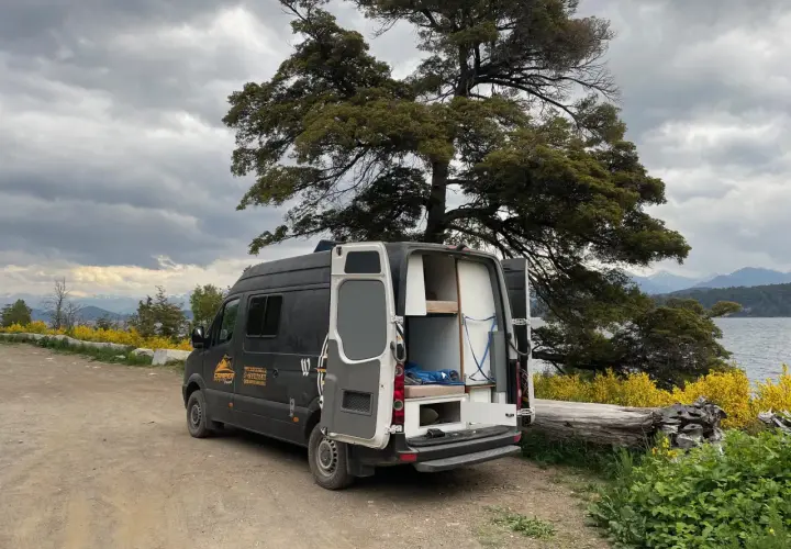 Un camping-car noir avec les portes arrière ouvertes, prêt pour un entretien, est garé près d'un lac sous un ciel nuageux.