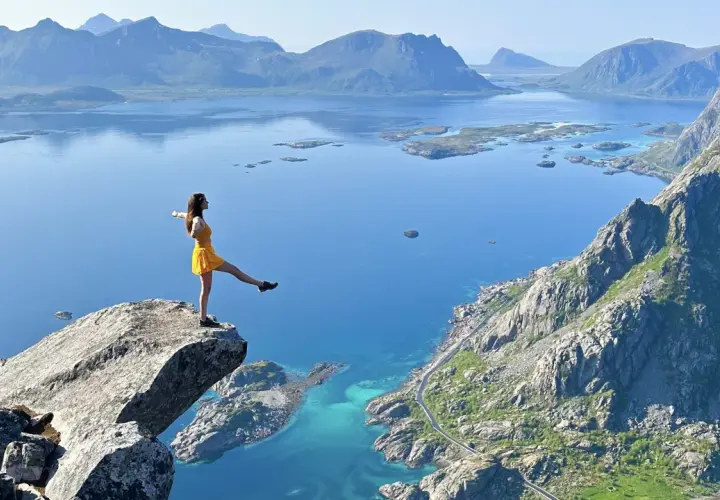 Une femme en robe jaune sur un promontoire rocheux, admirant les plus beaux endroits des îles Lofoten, avec des montagnes bleues et des eaux turquoise.
