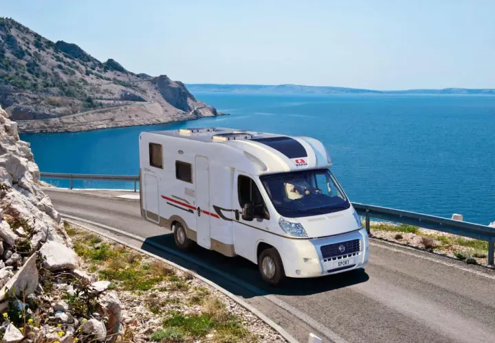 Un camping-car Adria roule sur une route côtière en Sardaigne, avec la mer bleue et des montagnes rocheuses en arrière-plan.