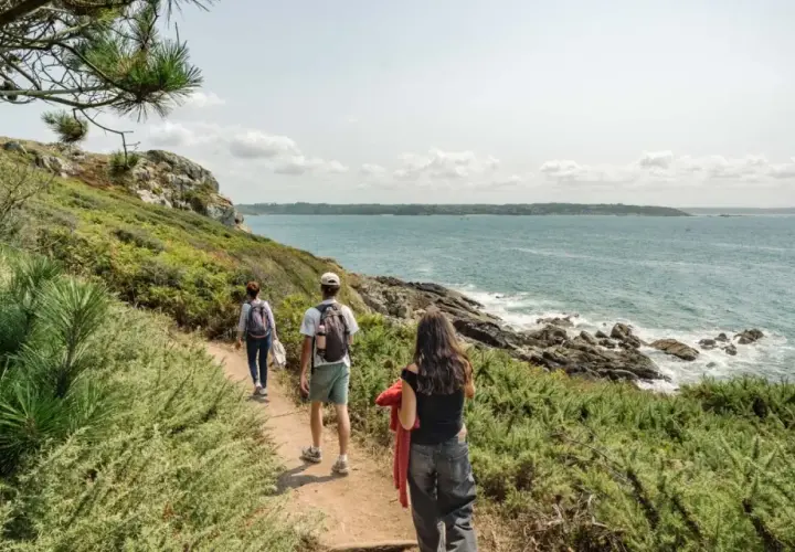 Trois randonneurs explorent le sentier des douaniers, longeant la côte rocheuse et la mer d'un bleu profond sous un ciel nuageux.