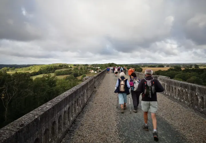 Un groupe de randonneurs, dont certains portent des sacs à dos et utilisent des bâtons de marche, traverse un ancien aqueduc en pierre sous un ciel couvert. Le paysage verdoyant s'étend à perte de vue.