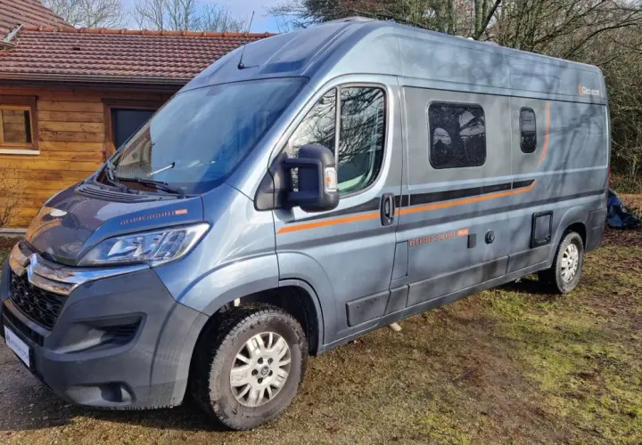 Un camping-car gris, modèle Globecar, est garé devant une maison en bois. Il est prêt pour une aventure avec son look d'Argus.