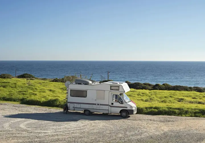 Camping-car blanc garé sur une aire gratuite en bord de mer en Bretagne. L'océan bleu s'étend à l'horizon, avec des champs verdoyants.