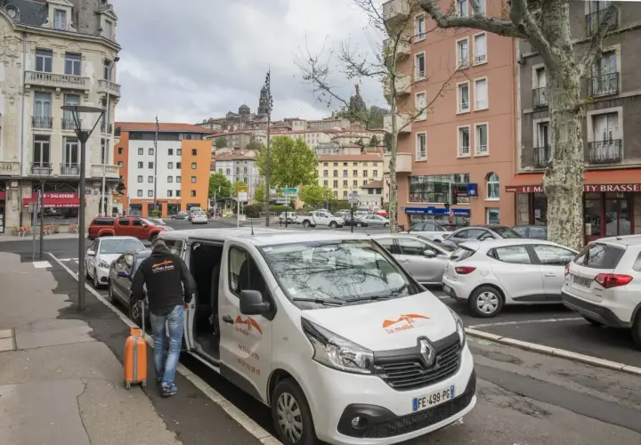 Un homme charge une valise orange dans un bus compostelle blanc. La ville s'étend en arrière-plan.