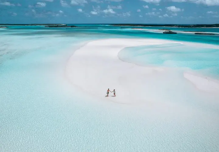 Un couple se tient la main sur une plage de sable blanc aux Bahamas. L'eau turquoise et les îlots au loin invitent à explorer que faire aux Bahamas.