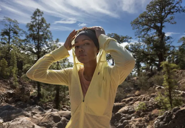 Une femme ajuste son bandeau et sa veste légère, parfaite pour savoir comment s'habiller pour une randonnée en été. Paysage rocheux et arbres en arrière-plan.