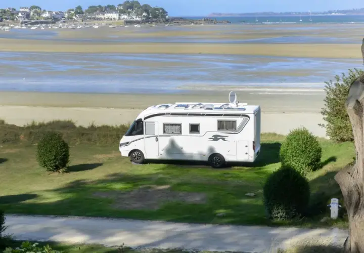 Vue d'une aire de camping-car avec sanitaire, face à une plage et un village côtier. Le camping-car blanc est garé sur l'herbe.