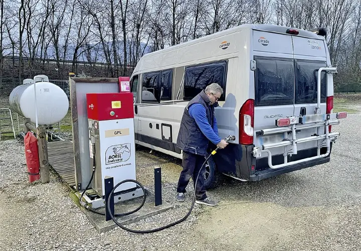 Un homme fait le plein de son camping-car à une installation GPL. Le véhicule est garé à côté d'une pompe à carburant rouge et blanche.