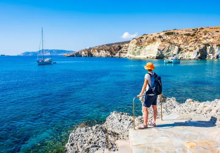 Un voyageur admire la mer Égée bleue et les falaises blanches, un voilier au loin. Le tourisme en Grèce offre des paysages magnifiques.
