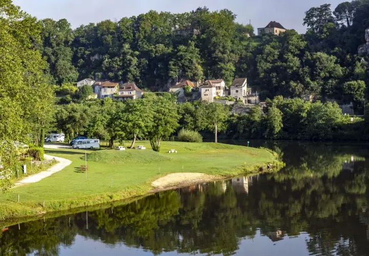Vue paisible d'un camping-car près de la rivière, avec des maisons pittoresques accrochées à la colline, rappelant le charme de Figeac et Cahors.