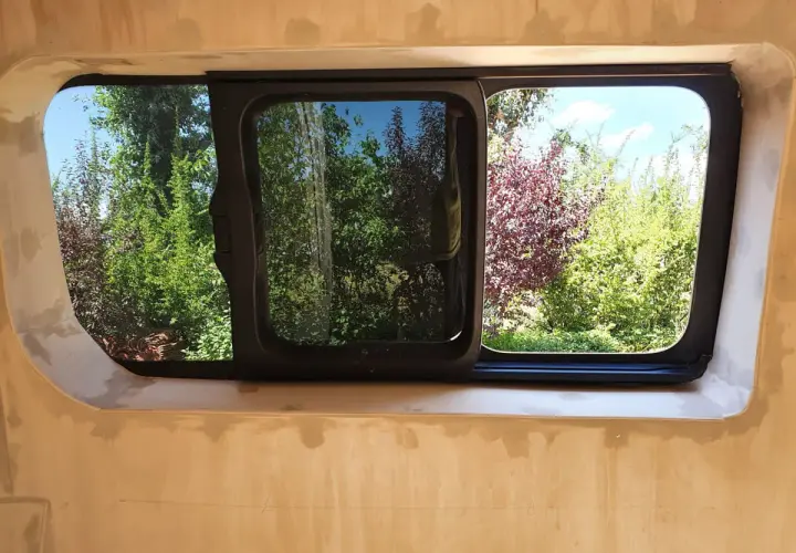 Fenêtre de fourgon aménagé, prête pour le contrôle technique. Vue sur un jardin verdoyant et fleuri.