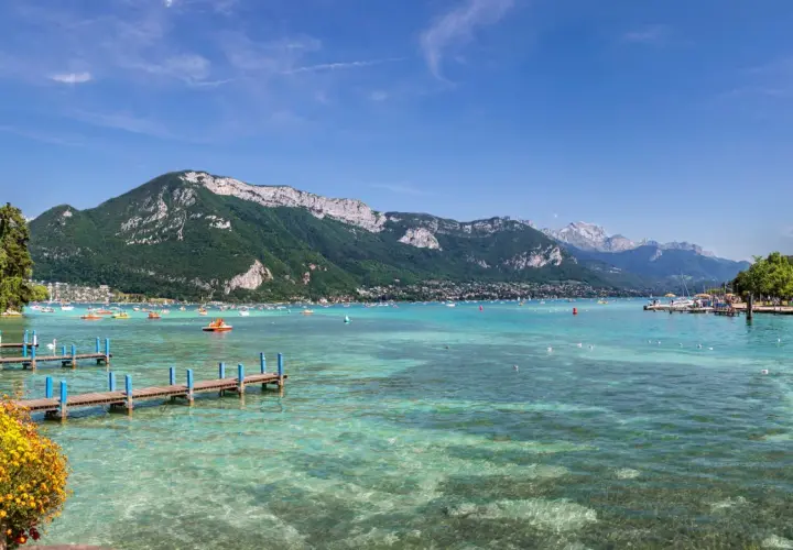 Vue panoramique du lac d'Annecy, avec des montagnes verdoyantes en arrière-plan. Des bateaux et des pédalos animent les eaux turquoise. Un ponton fleuri invite à la promenade.