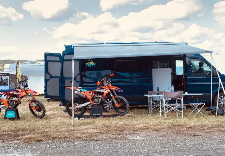Un fourgon aménagé avec soute moto, prêt pour l'aventure. Deux motos de cross orange attendent devant, sous un ciel nuageux.