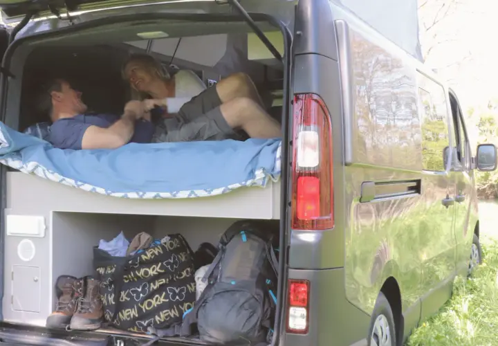 Couple joyeux dans un camping-car lit transversal. Sacs et chaussures de randonnée rangés en dessous.