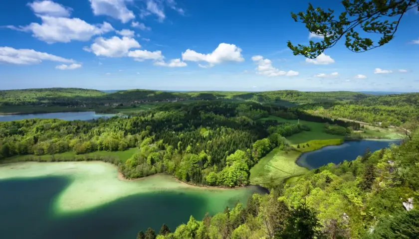 Paysage verdoyant du Jura avec des lacs aux eaux claires, des forêts denses et un ciel bleu parsemé de nuages blancs.