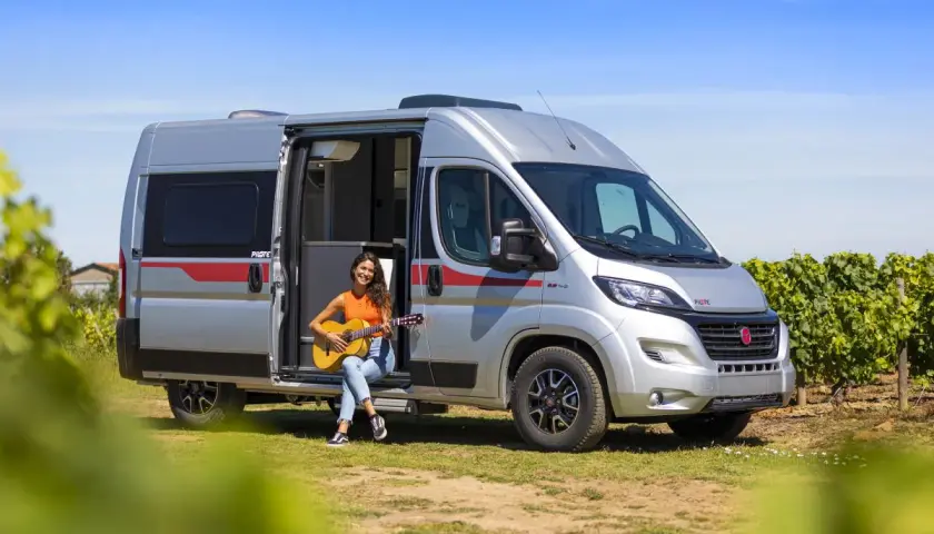 Une jeune femme joue de la guitare assise à l'entrée d'un camping-car aménagé, un type de camping car idéal pour les escapades ensoleillées.