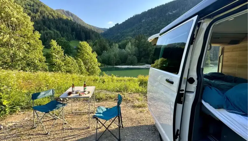 Un coin de paradis pour la Bourgogne en camping-car : table dressée, chaises bleues, lac paisible et montagnes verdoyantes.