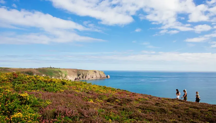 Trois femmes profitent d'un trek en Bretagne, longeant une côte rocheuse et fleurie, sous un ciel bleu parsemé de nuages.