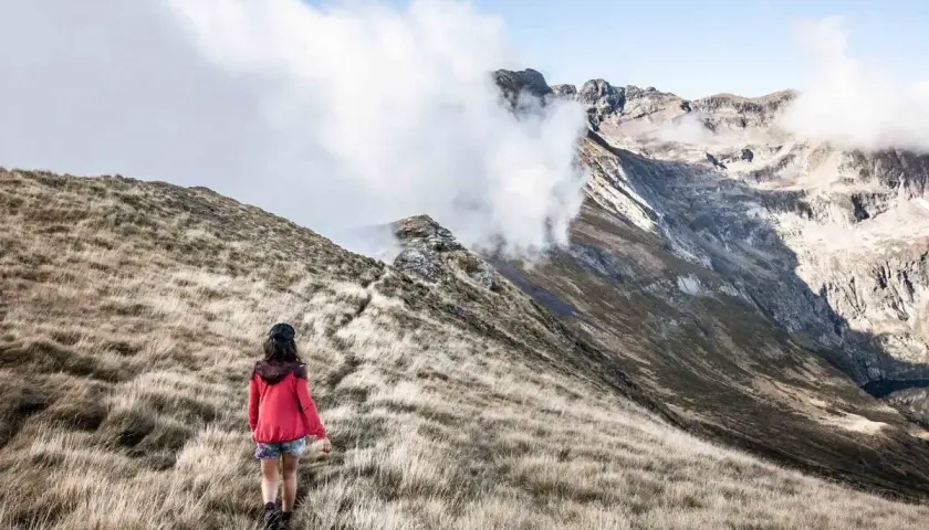 Une randonneuse explore un sentier herbeux, s'approchant des majestueuses montagnes du tour du Biros, enveloppées de nuages.