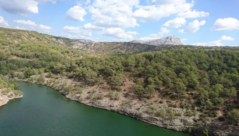 Vue panoramique du lac Zola, entouré de collines verdoyantes et rocheuses sous un ciel bleu parsemé de nuages blancs.