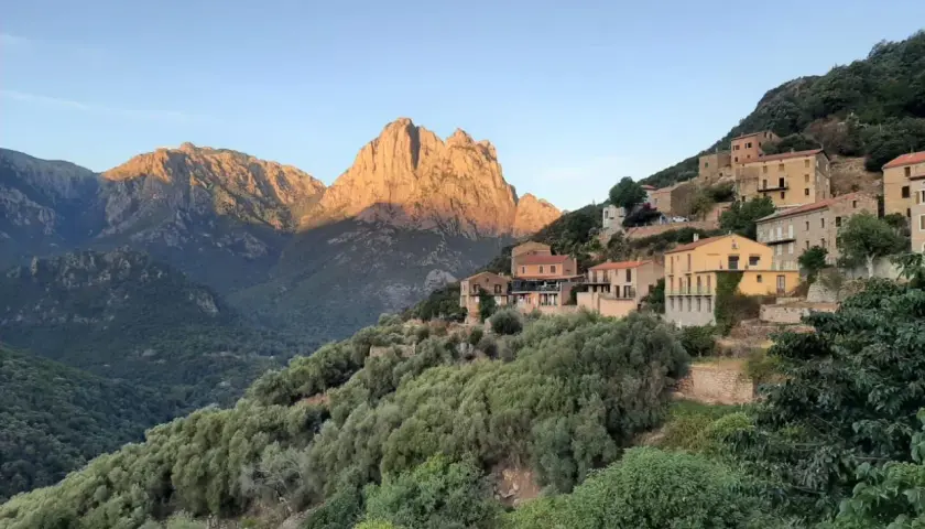 Village perché sur une colline, baigné par le soleil couchant. Les montagnes du nord, d'une difficulté certaine, se dressent en arrière-plan.
