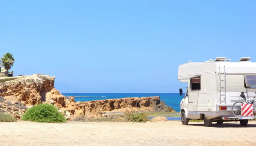 Camping-car blanc stationné sur une plage de sable en Espagne, avec la mer bleue et des falaises rocheuses en arrière-plan. Une aire de camping-car espagne gratuite idéale pour les vacances.