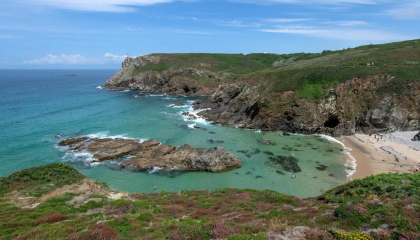 Vue panoramique de la pointe du Van, avec ses falaises verdoyantes, ses eaux turquoise et sa petite plage. Quelques baigneurs profitent du soleil.
