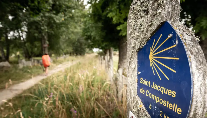Panneau Saint Jacques de Compostelle sur le chemin, indiquant la direction de Conques. Un pèlerin s'éloigne sur le sentier.