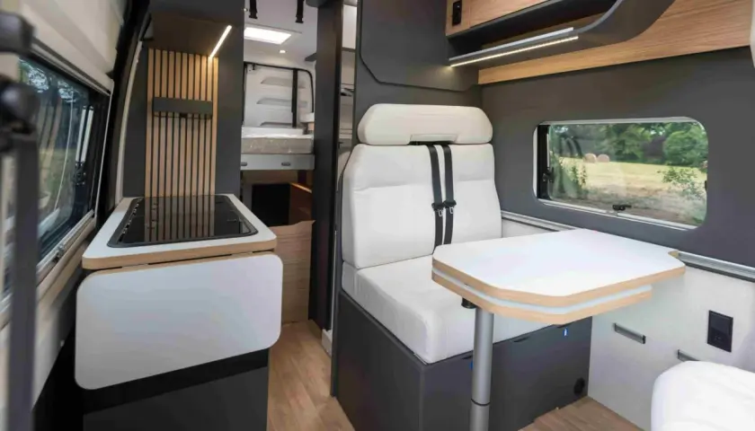 Intérieur d'un camping-car Bavaria : banquette confortable, table escamotable, plaque de cuisson et lit visible au fond.