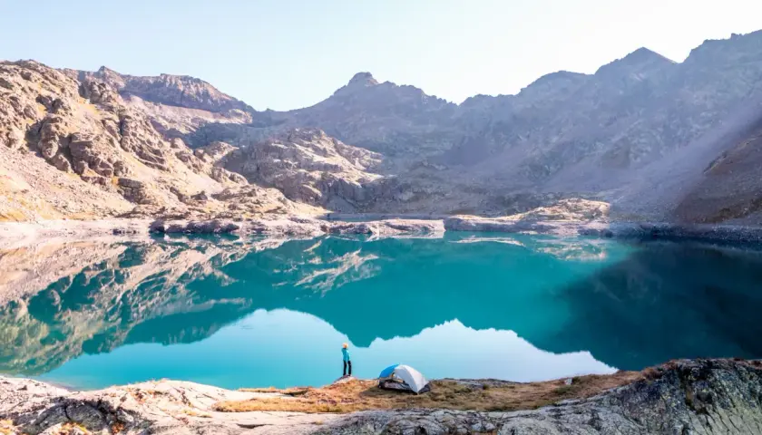 Un randonneur admire un lac turquoise aux reflets parfaits dans le **parc du Mercantour**. Une tente est installée sur la rive rocheuse.
