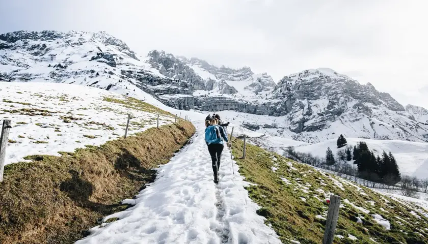 Une randonnée ce week-end dans un paysage montagneux enneigé. Des randonneurs suivent un sentier bordé de clôtures en bois, avec des sommets imposants en arrière-plan.