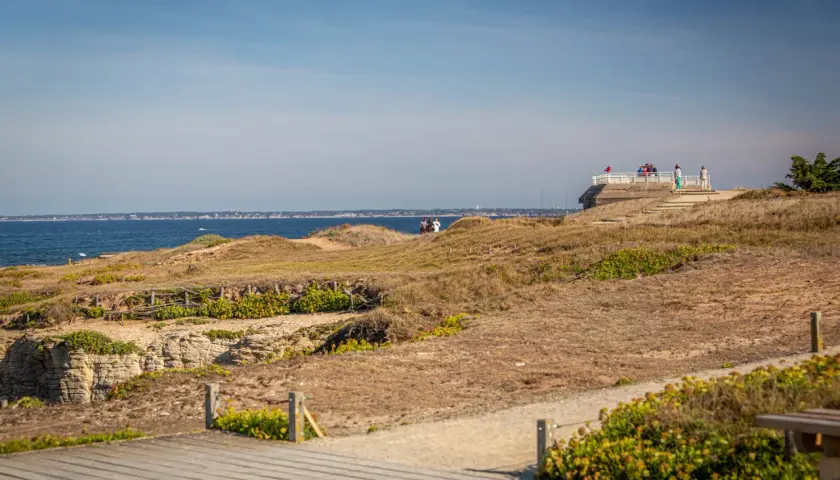 Vue panoramique sur la côte sauvage de la pointe Saint-Gildas, avec des falaises rocheuses, des dunes et la mer bleue. Des promeneurs profitent du paysage.
