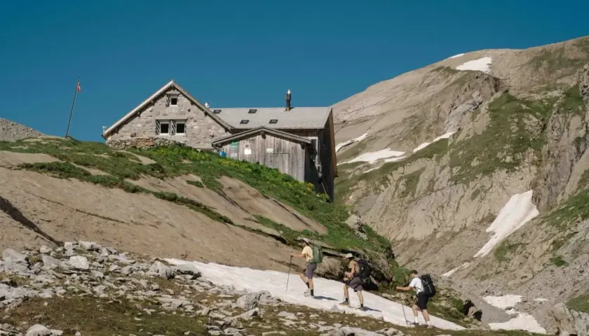 Trois randonneurs approchent d'une **cabane refuge** en pierre et bois, perchée sur une pente rocheuse et herbeuse, sous un ciel bleu intense.