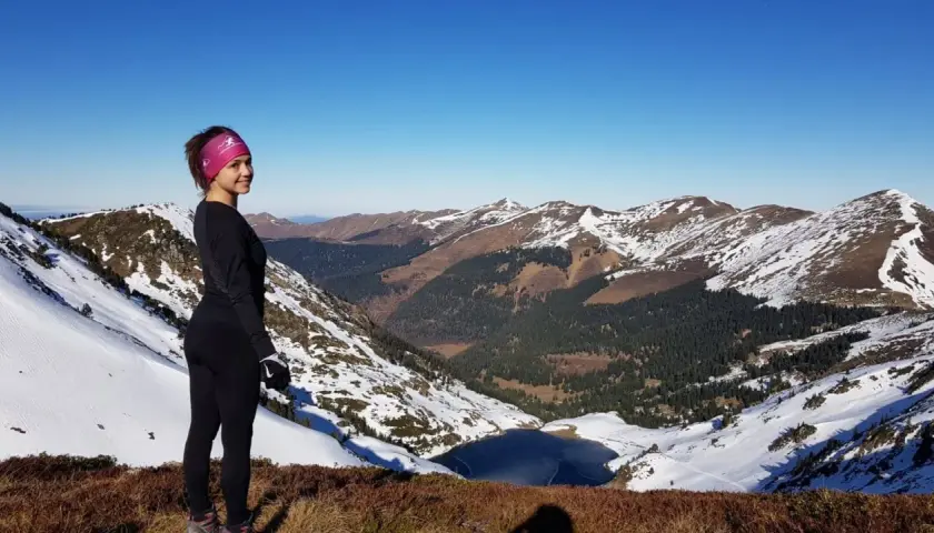 Vue panoramique depuis le pic du lion. Une femme souriante, en tenue de sport, admire le paysage montagneux enneigé et un lac gelé.