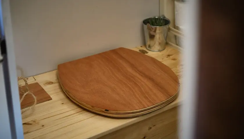 Un siège en bois pour toilettes sèches de camping-car, avec un petit seau de plantes et du papier toilette à proximité.