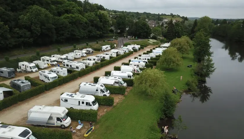 Vue aérienne d'une aire de camping-car gratuite en bord de mer normandie, avec de nombreux véhicules stationnés près d'une rivière bordée d'arbres.