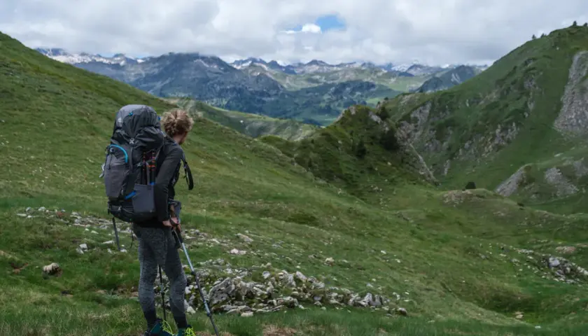 Une randonneuse admire le panorama des Pyrénées, prête pour son trek. Le soleil perce les nuages sur les sommets enneigés.