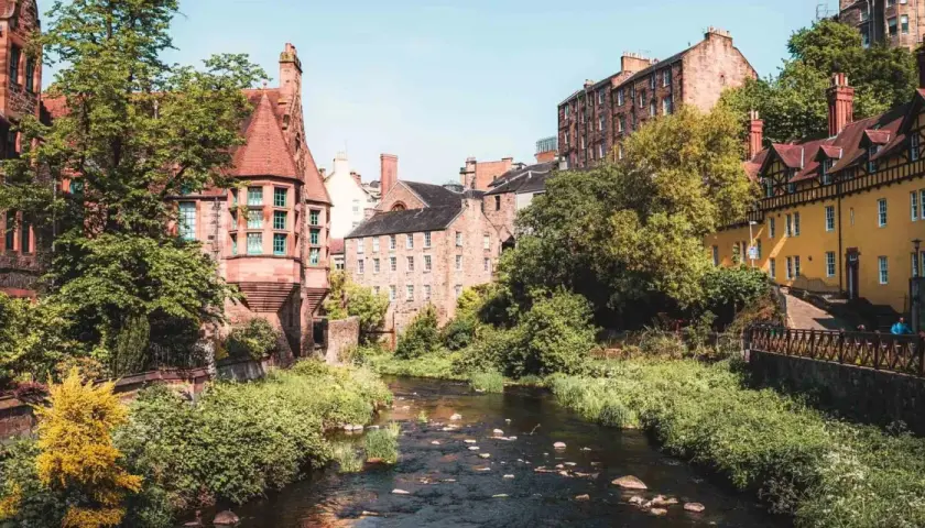 Un charmant ruisseau serpente à travers des bâtiments historiques en grès et une maison jaune vif. Idéal pour savoir que faire à Édimbourg.