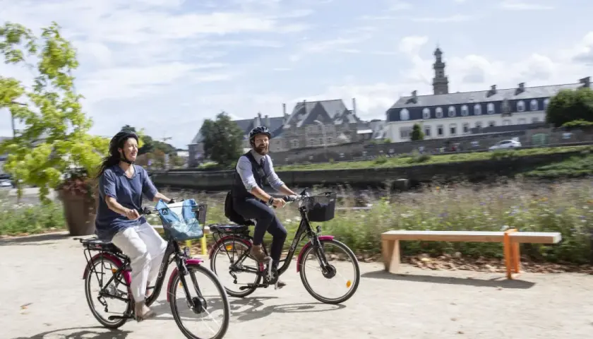 Couple à vélo profitant d'une balade ensoleillée le long de la rivière, avec les bâtiments historiques de Tréguier Lannion en arrière-plan.