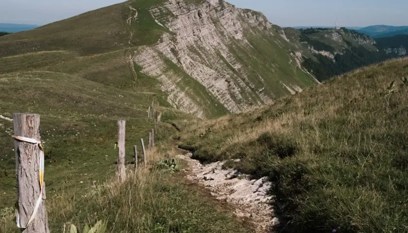 Sentier rocailleux serpentant dans l'herbe haute, menant vers des montagnes escarpées. Une clôture rudimentaire marque le chemin de cette échappée jurassienne.