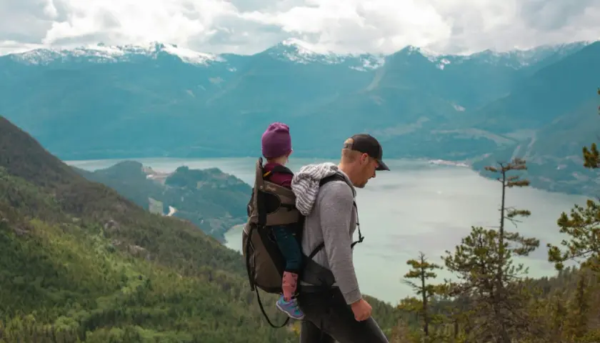 Un père porte son enfant dans un porte-bébé lors d'un week-end randonnée, avec des montagnes enneigées en arrière-plan.