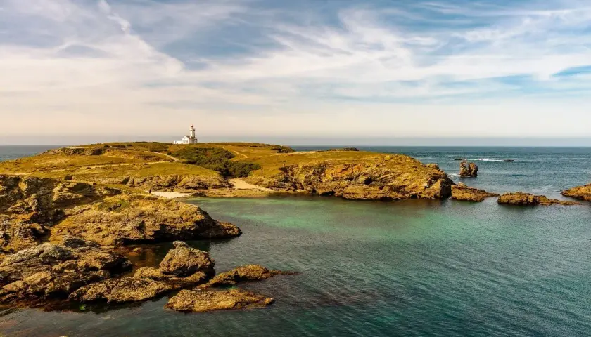 Phare blanc sur une colline verdoyante d'une île bretonne, avec des rochers dorés et une mer turquoise sous un ciel nuageux.
